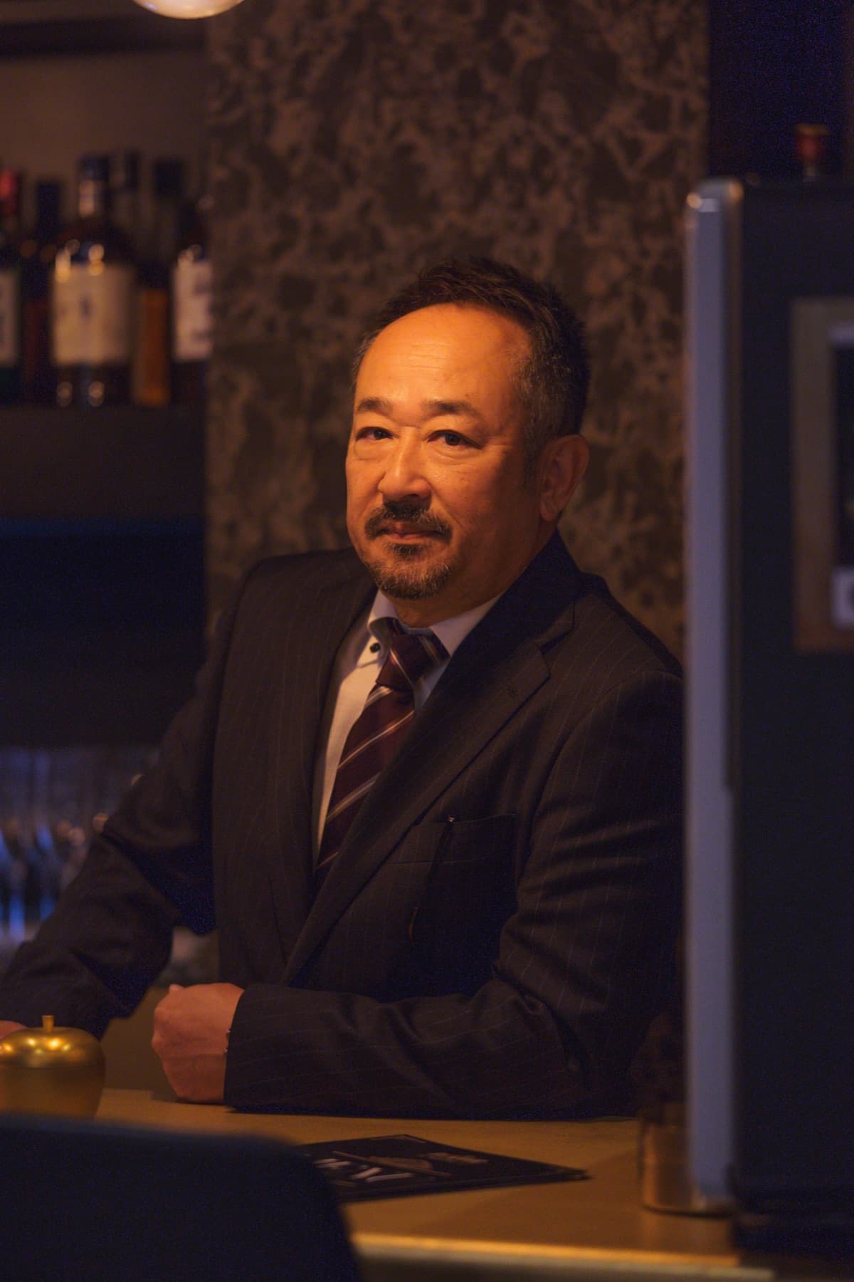 The Backdoor's owner at the counter — suit, striped tie, whisky shelves softly out of focus behind him.