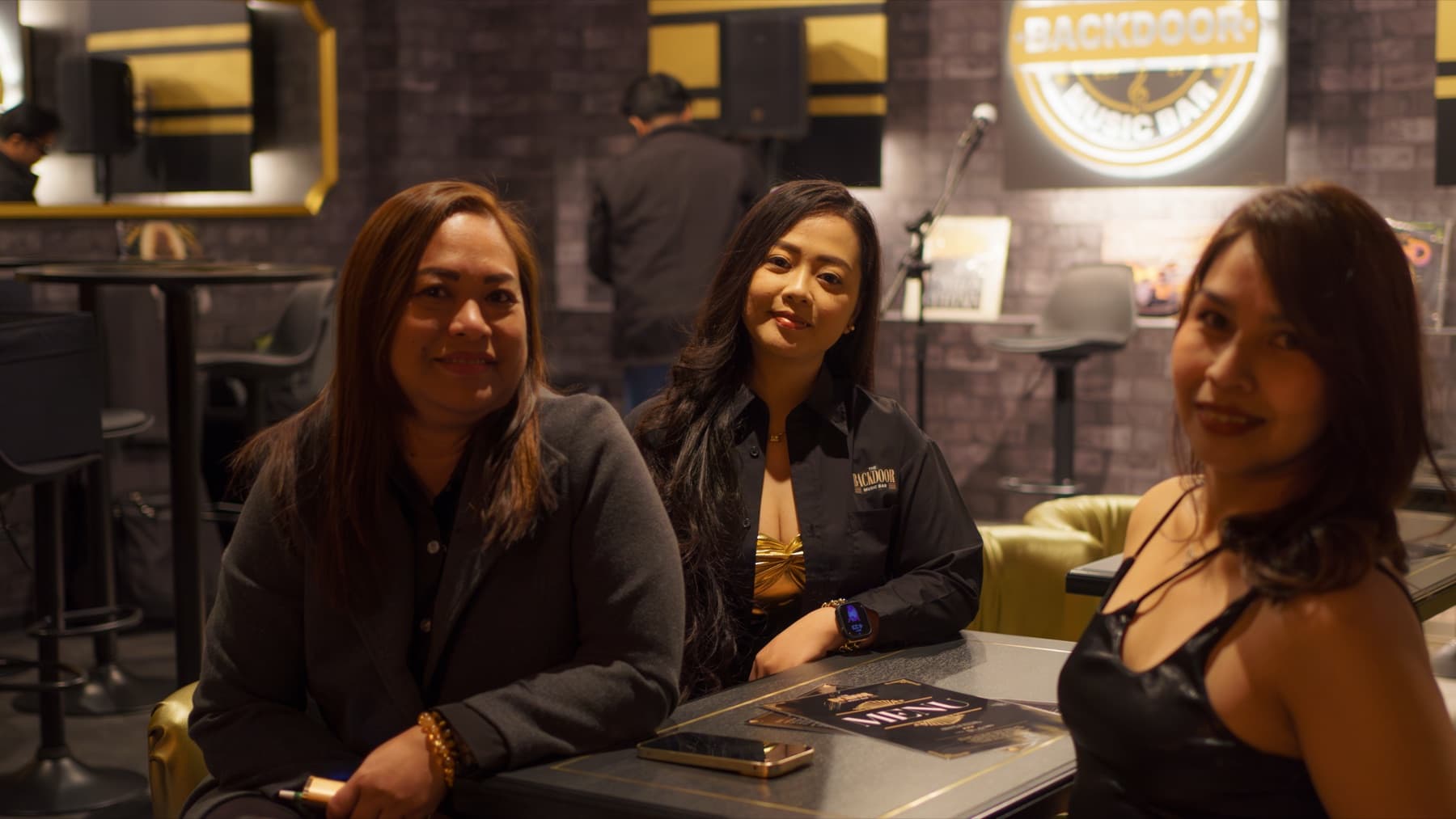 Three guests at a booth table in the Backdoor, leather seating, the illuminated Backdoor sign behind them.