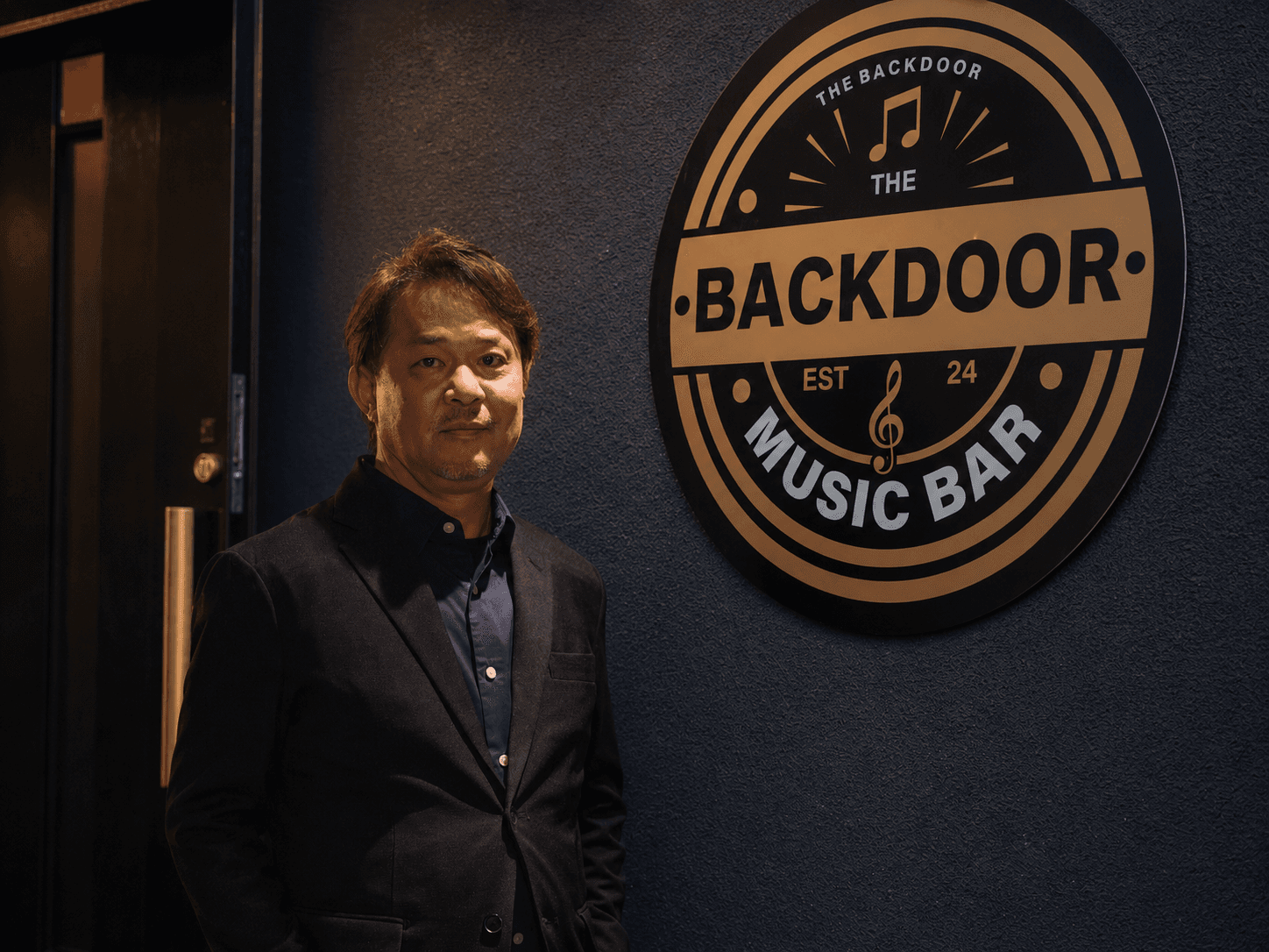Entrance to The Backdoor . the owner standing beside the large brass Backdoor badge mounted on the exterior wall, a floral arrangement to the right.