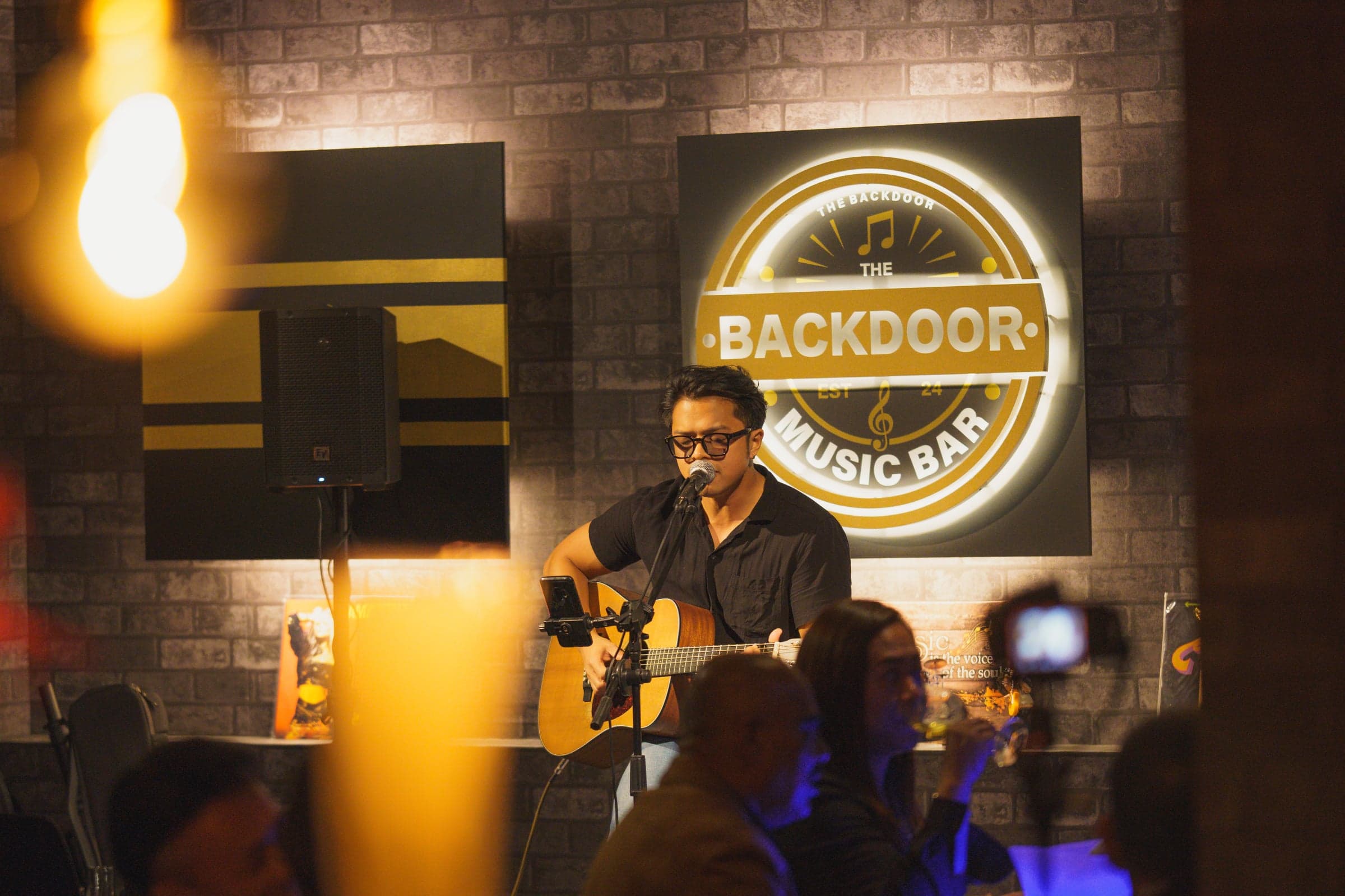 Vocalist in a black slip dress on the Backdoor stage, in front of the illuminated Backdoor logo, holding a microphone.